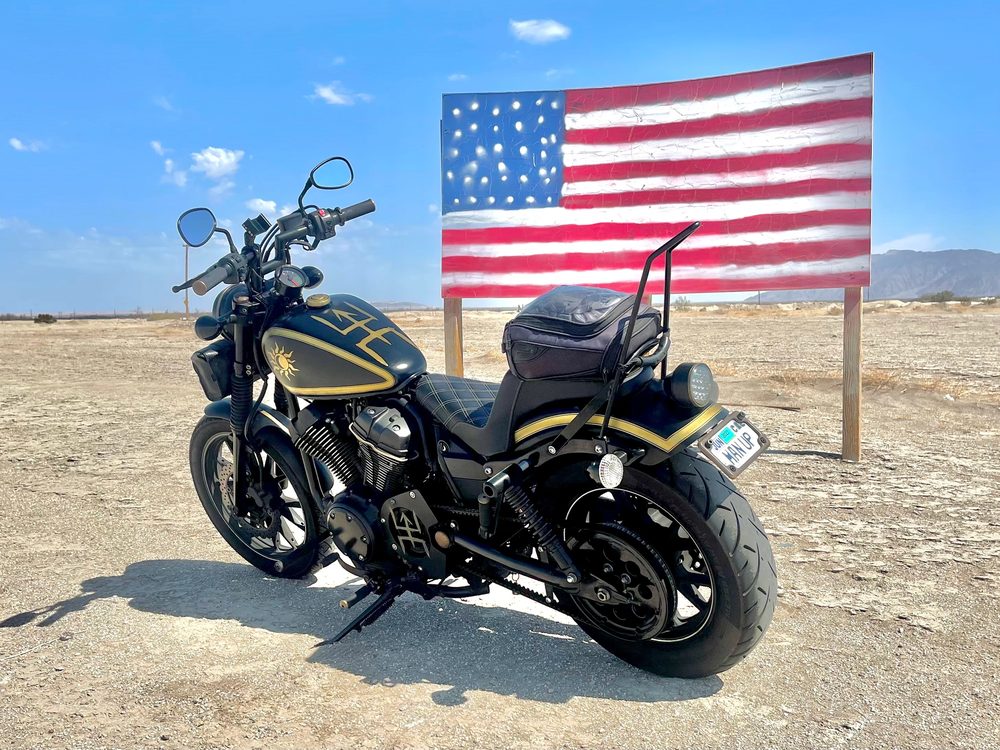 Dennis's motorcycle with the Man-UP! Life Coaching logo, parked by an American flag in the desert